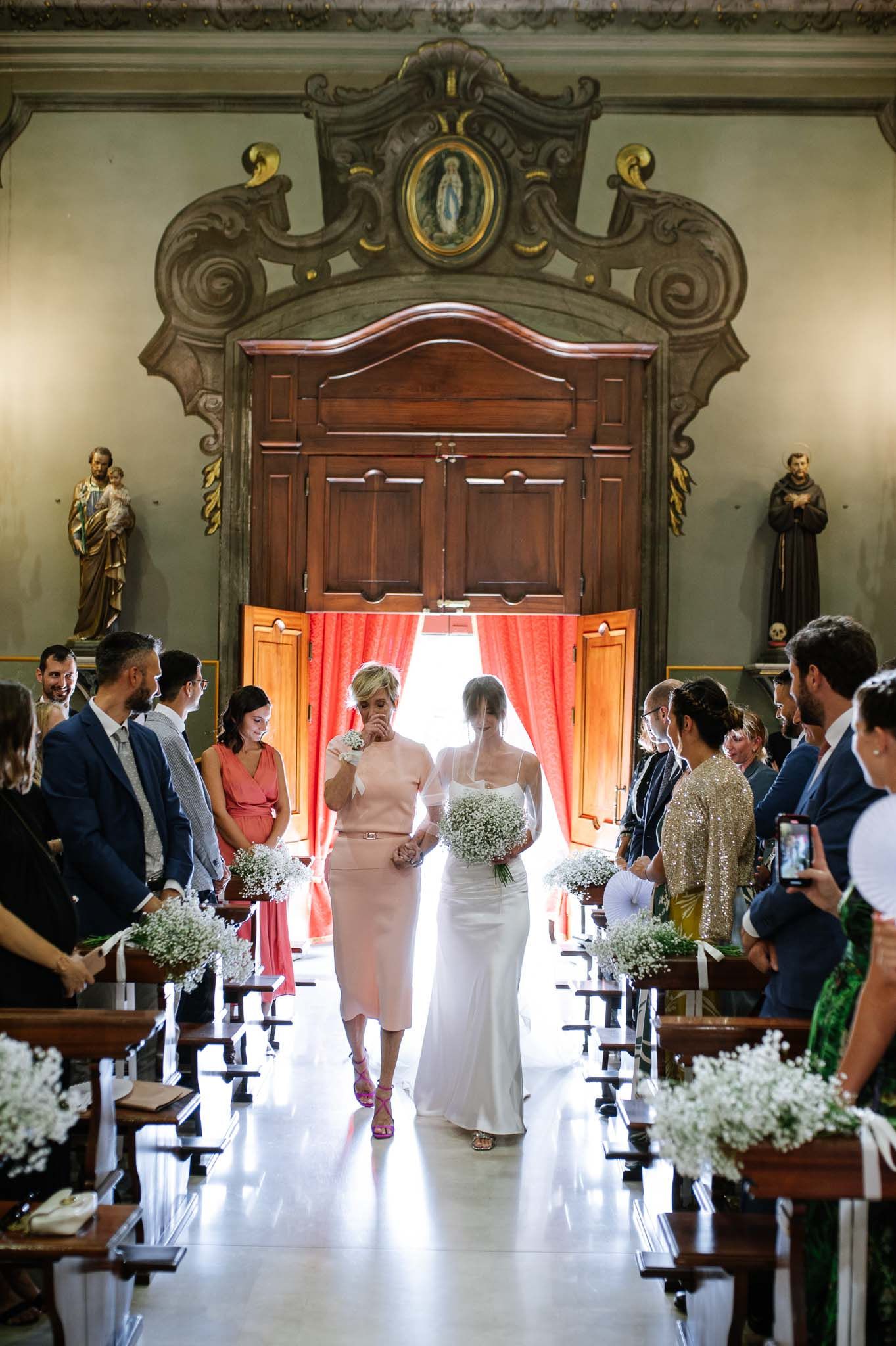 A bride walks down a church aisle decorated with baby's breath under ornate architectural details.