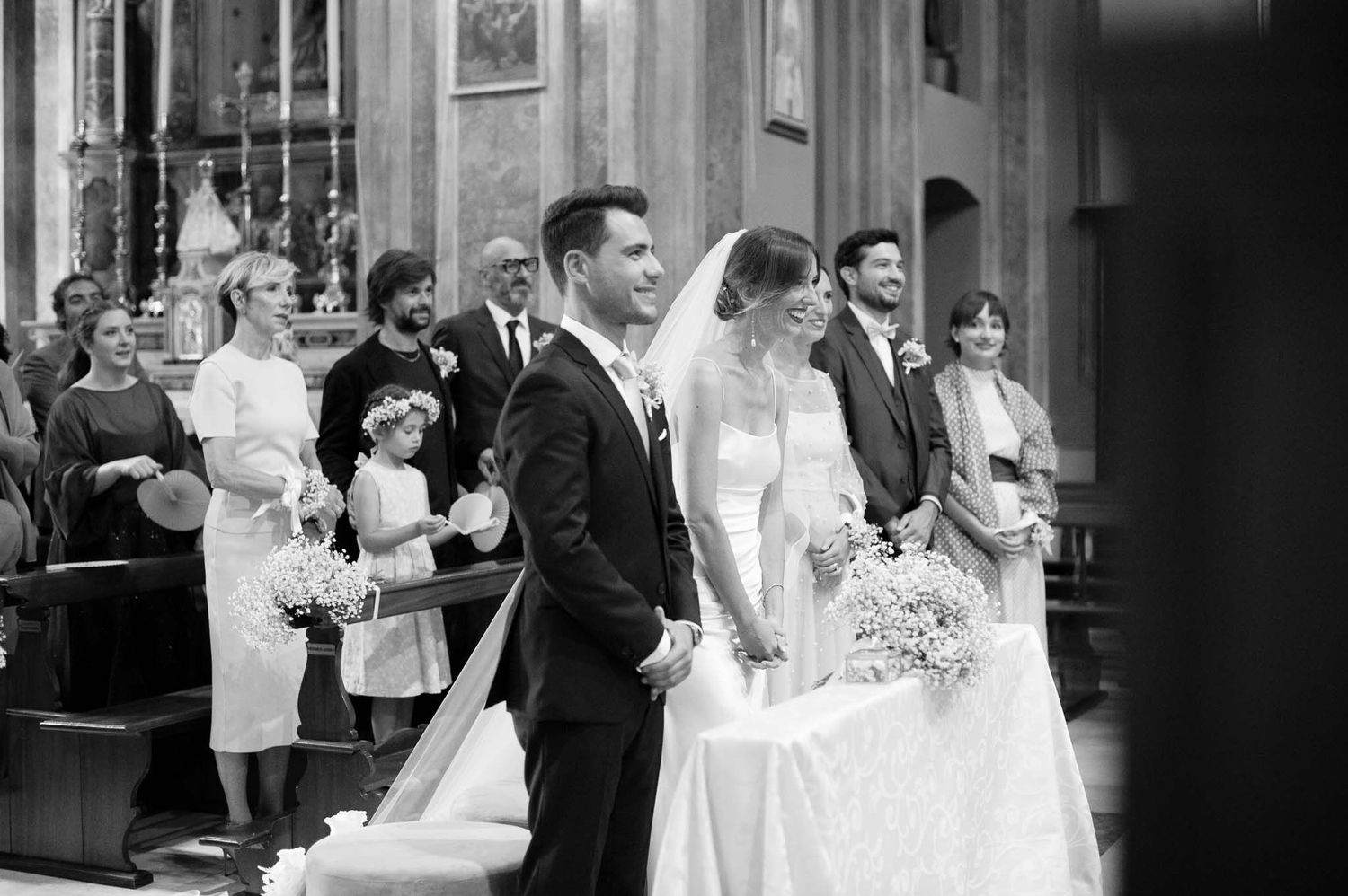 Wedding ceremony in a grand church with guests gathered around the decorated altar in black and white.