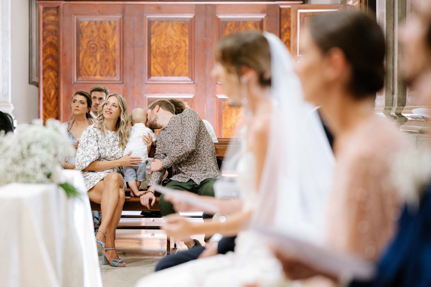 Wedding guests seated in wooden church pews during ceremony with soft natural lighting.