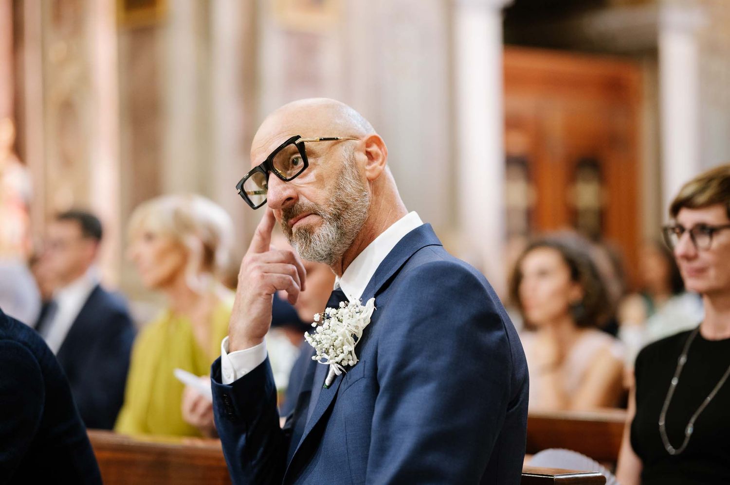 Wedding guest wearing glasses and boutonniere observing ceremony in church pew.