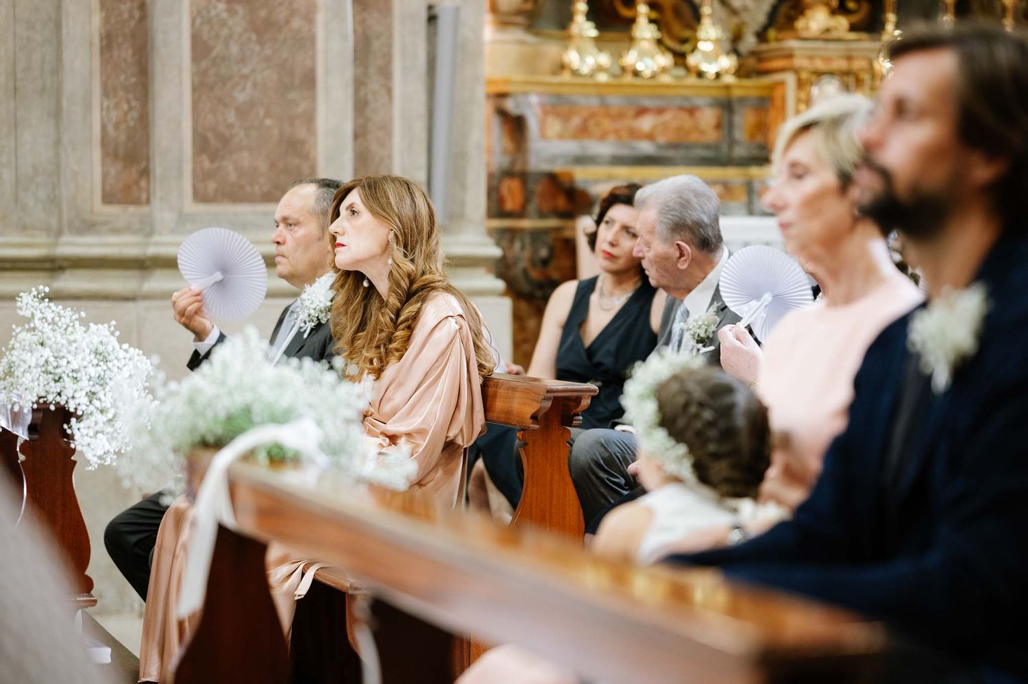 Wedding attendees seated in wooden pews during ceremony in ornate church interior.