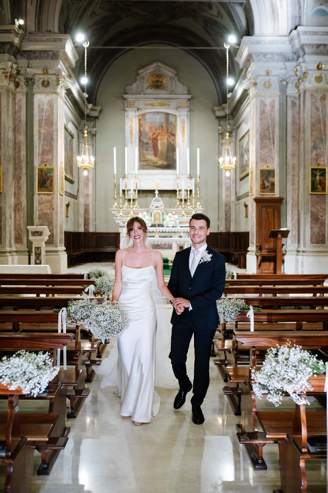 Couple walks down the aisle of an ornate Italian church decorated with white floral arrangements.