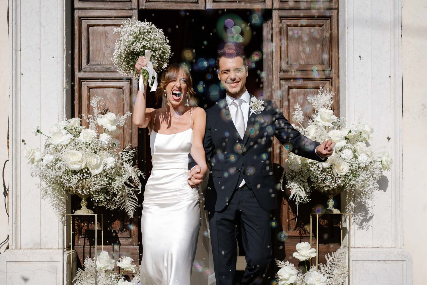 Newlyweds celebrate joyfully under confetti outside church doors decorated with white floral arrangements.
