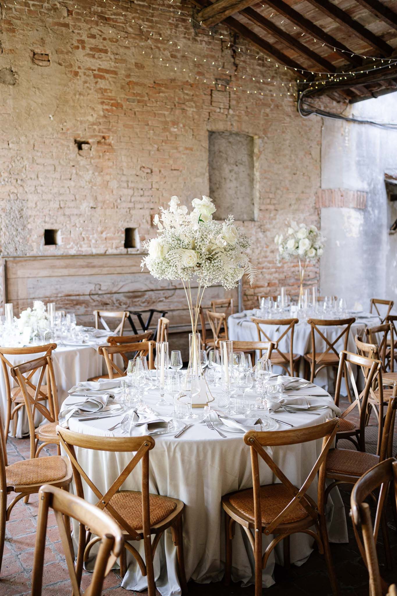 Elegant rustic wedding reception table with white floral centerpiece and cross-back chairs in brick venue.