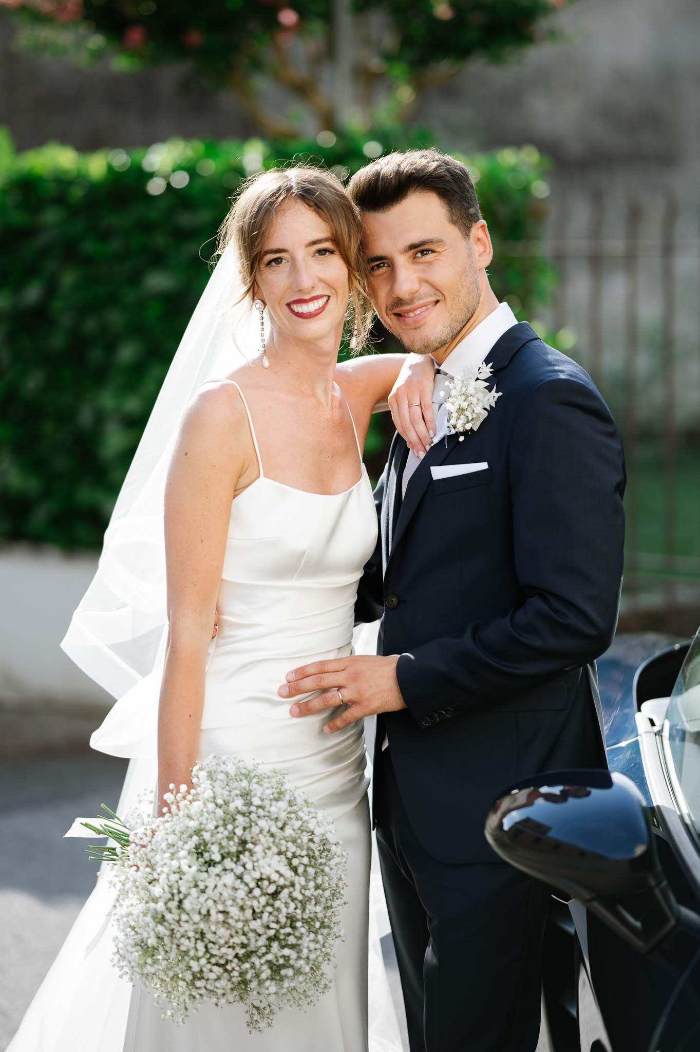 Wedding couple poses together outdoors with luxury car in background.