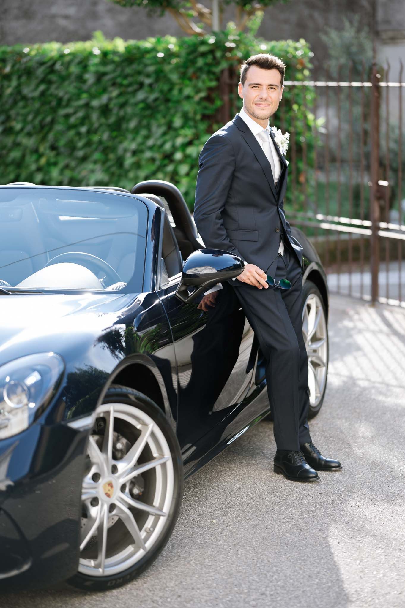 Groom in dark suit poses next to luxury sports car outdoors.