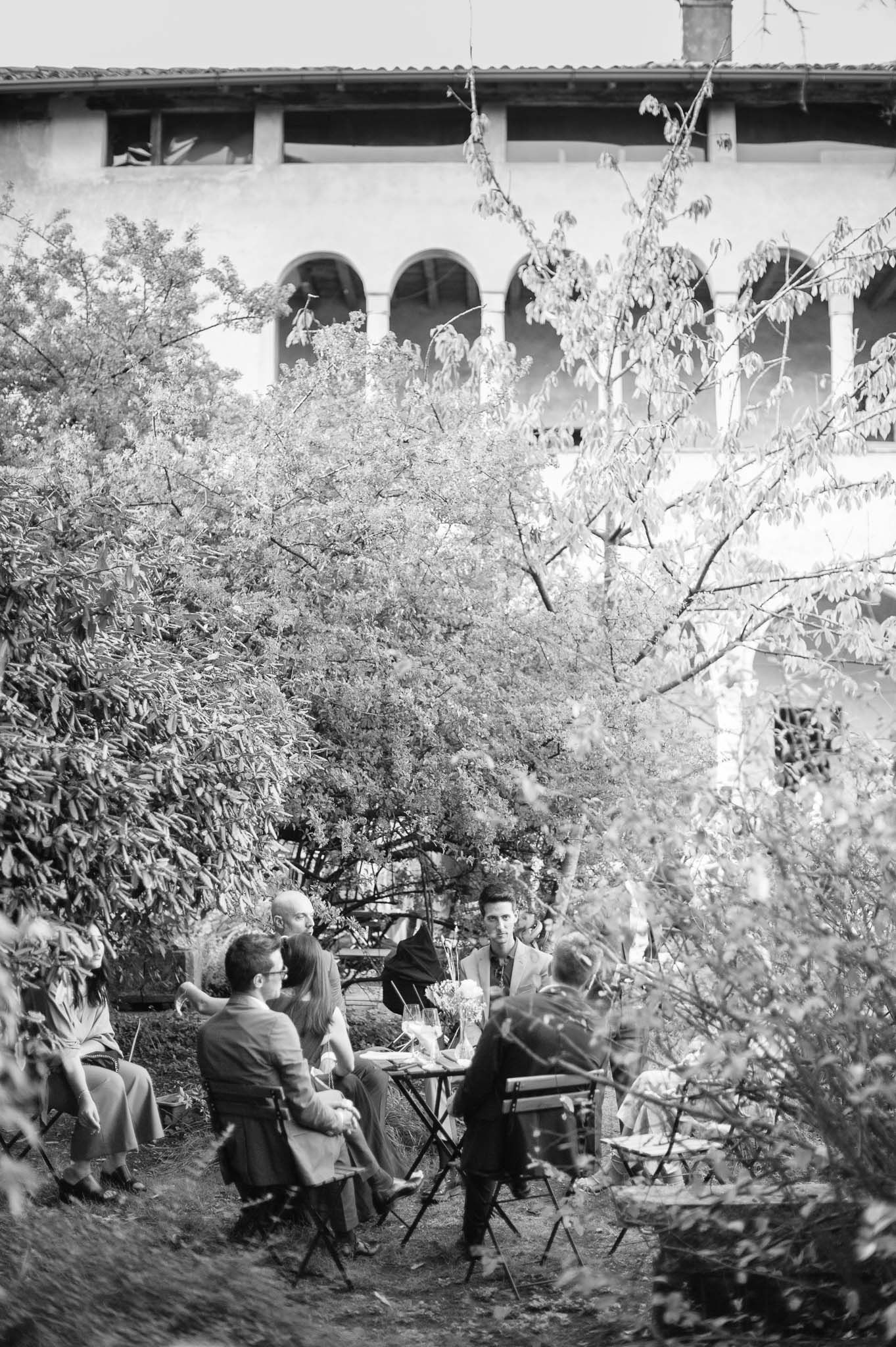 A vintage black and white photo showing people dining outdoors under arched architecture surrounded by foliage.
