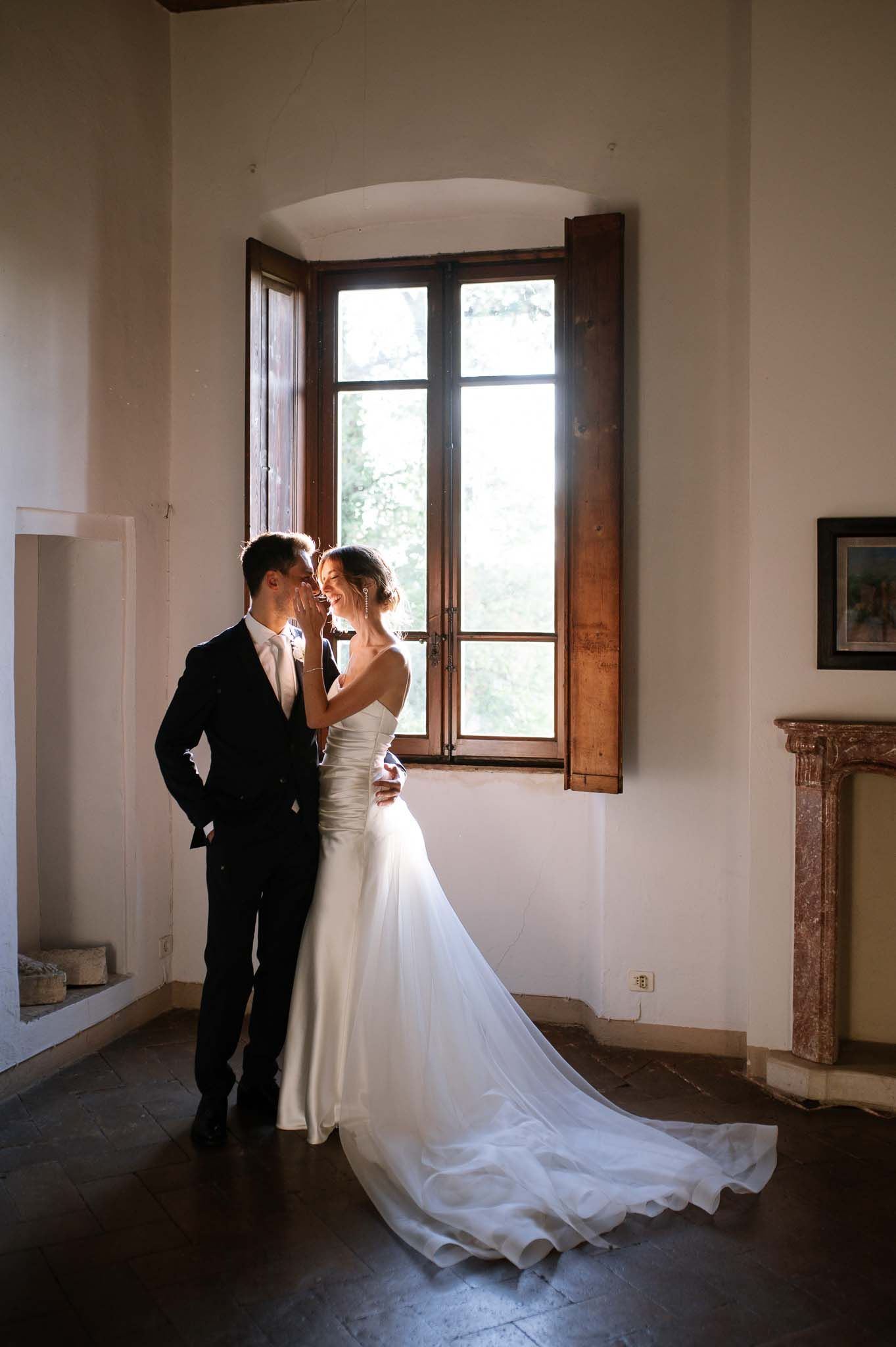 Romantic portrait of newlyweds by a rustic window with natural light streaming in.