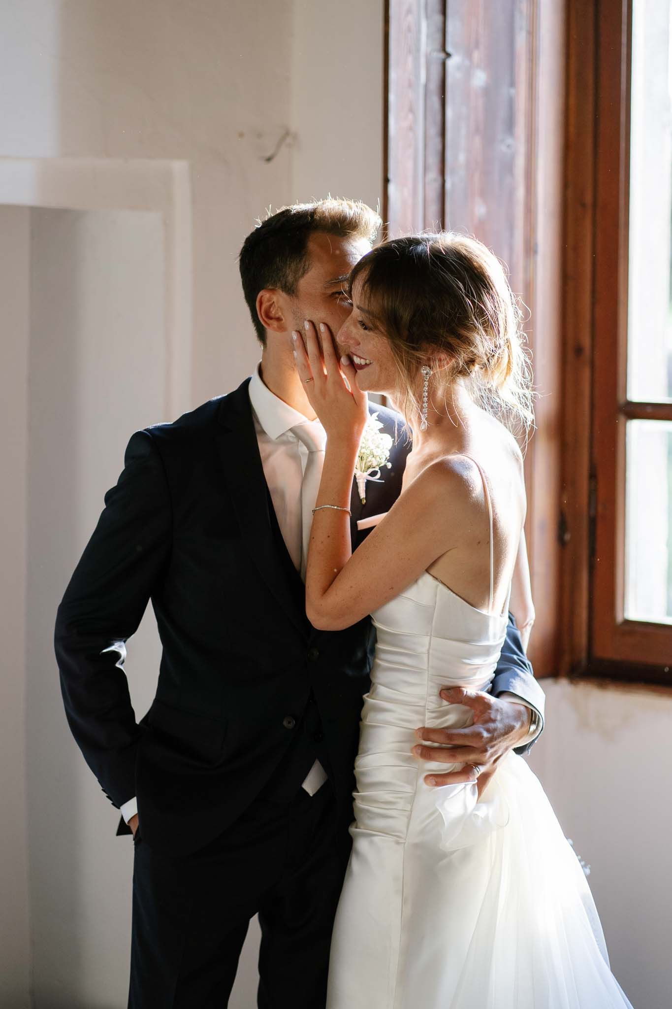 A tender moment between a couple in wedding attire sharing an intimate embrace near a window.