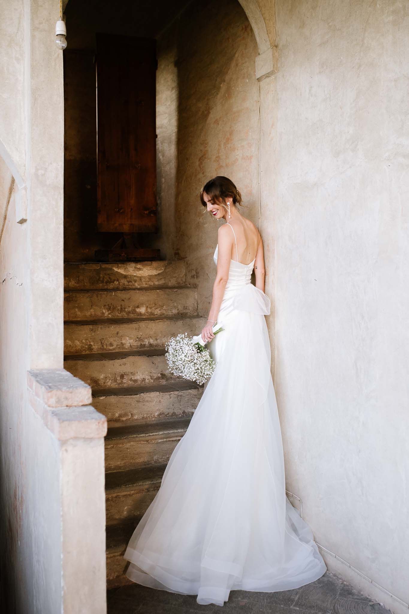 A bride in an elegant white gown holds a bouquet while standing on rustic stone stairs.