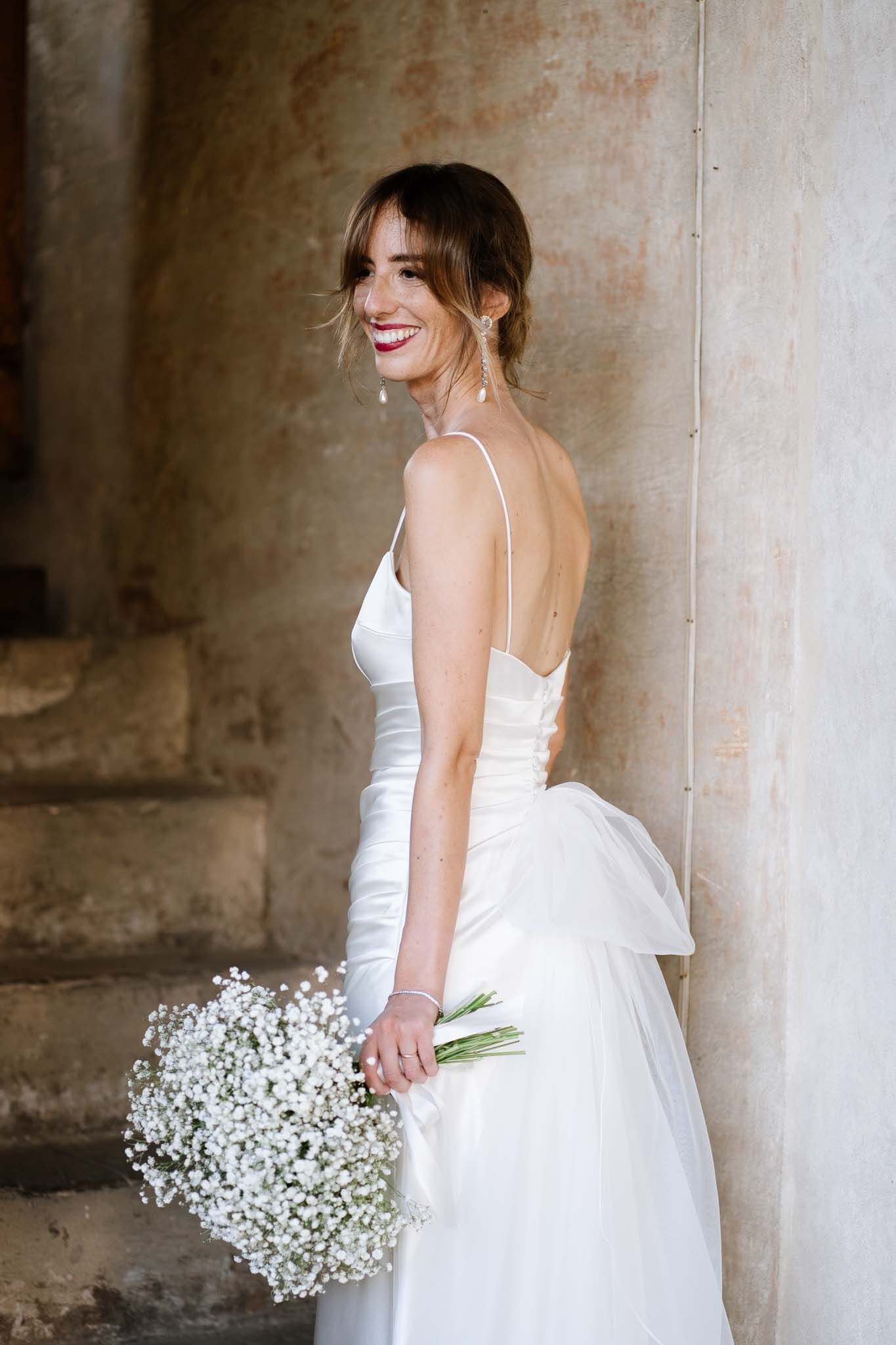 A joyful bride holding a baby's breath bouquet poses in a flowing white dress against a stone wall.