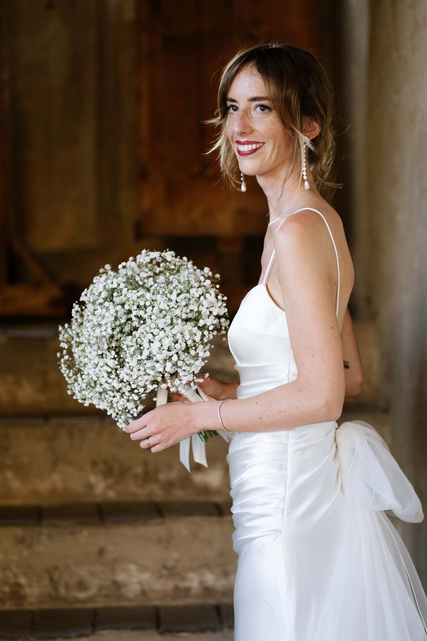 A radiant bride smiles while holding a white baby's breath bouquet on stone steps.