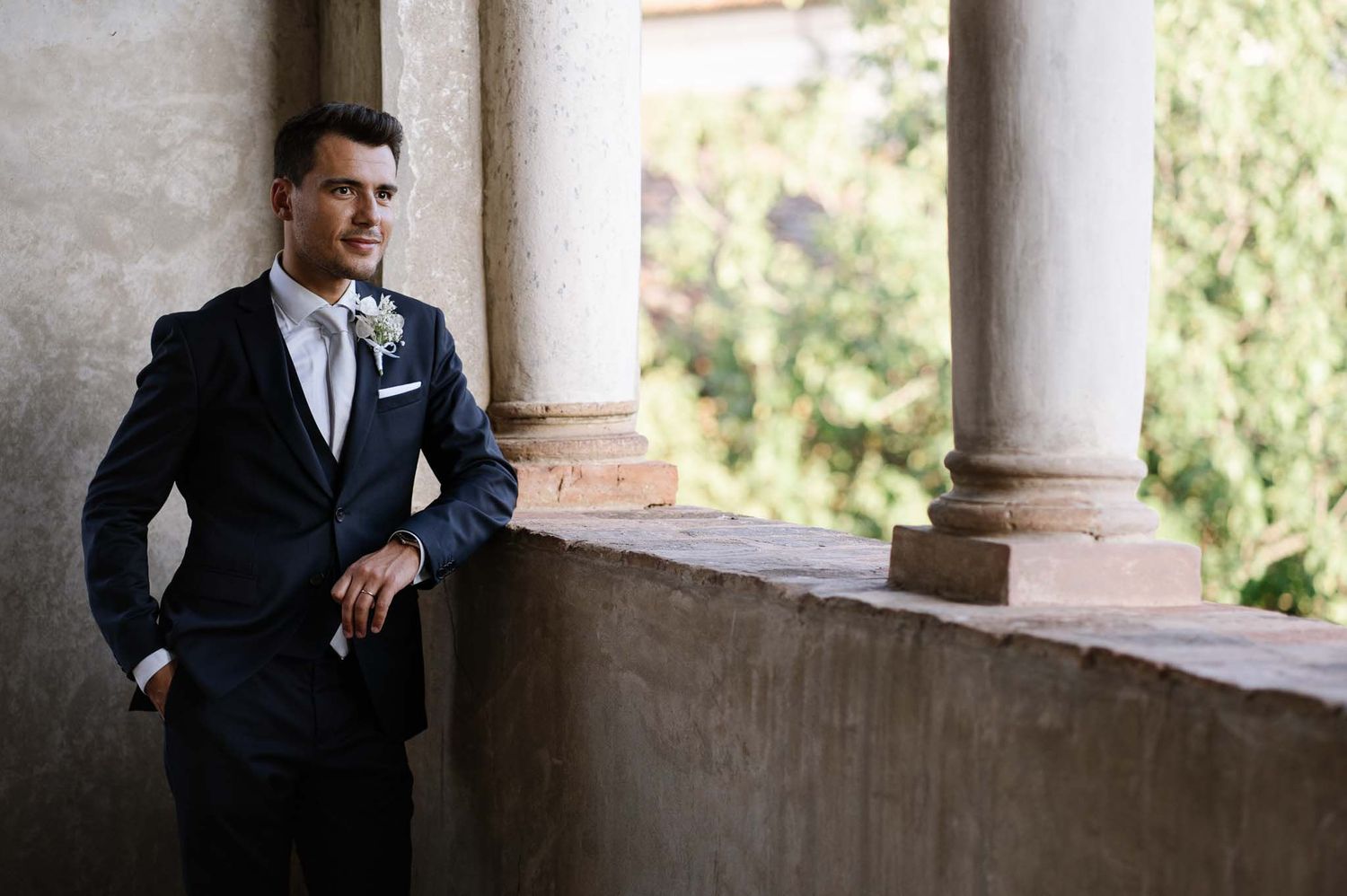A groom in a dark suit poses elegantly against stone columns with natural lighting.