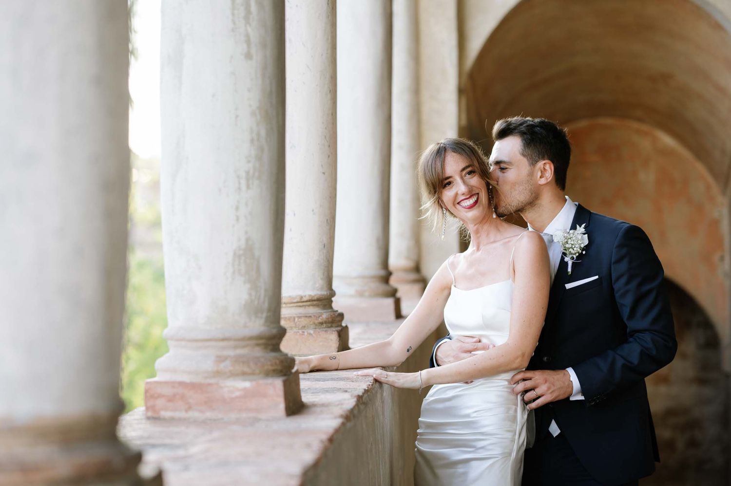 Elegant wedding couple poses in a sunlit stone archway with classic architecture.