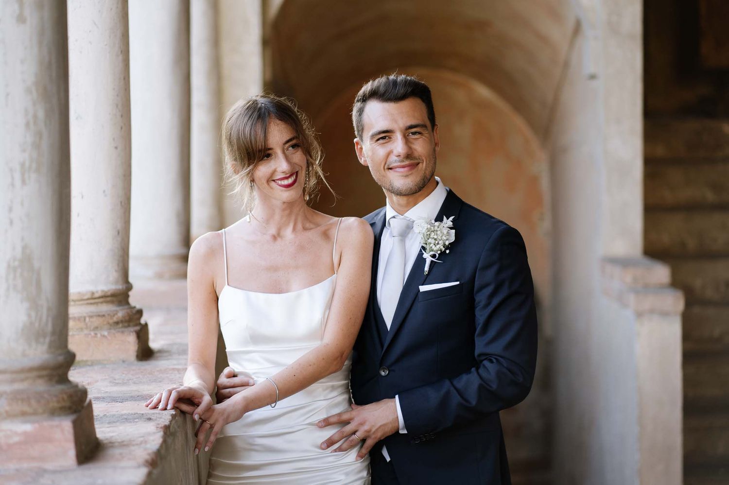 Happy newlyweds pose together against classical stone pillars during their wedding.