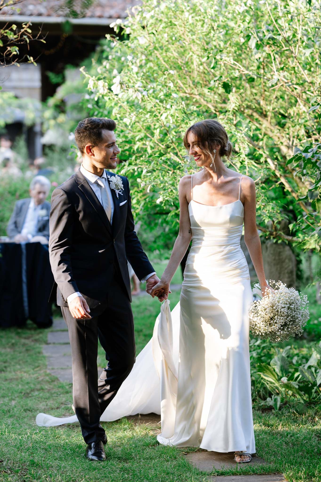 Bride in silk gown and groom in tuxedo walk through garden during wedding celebration.