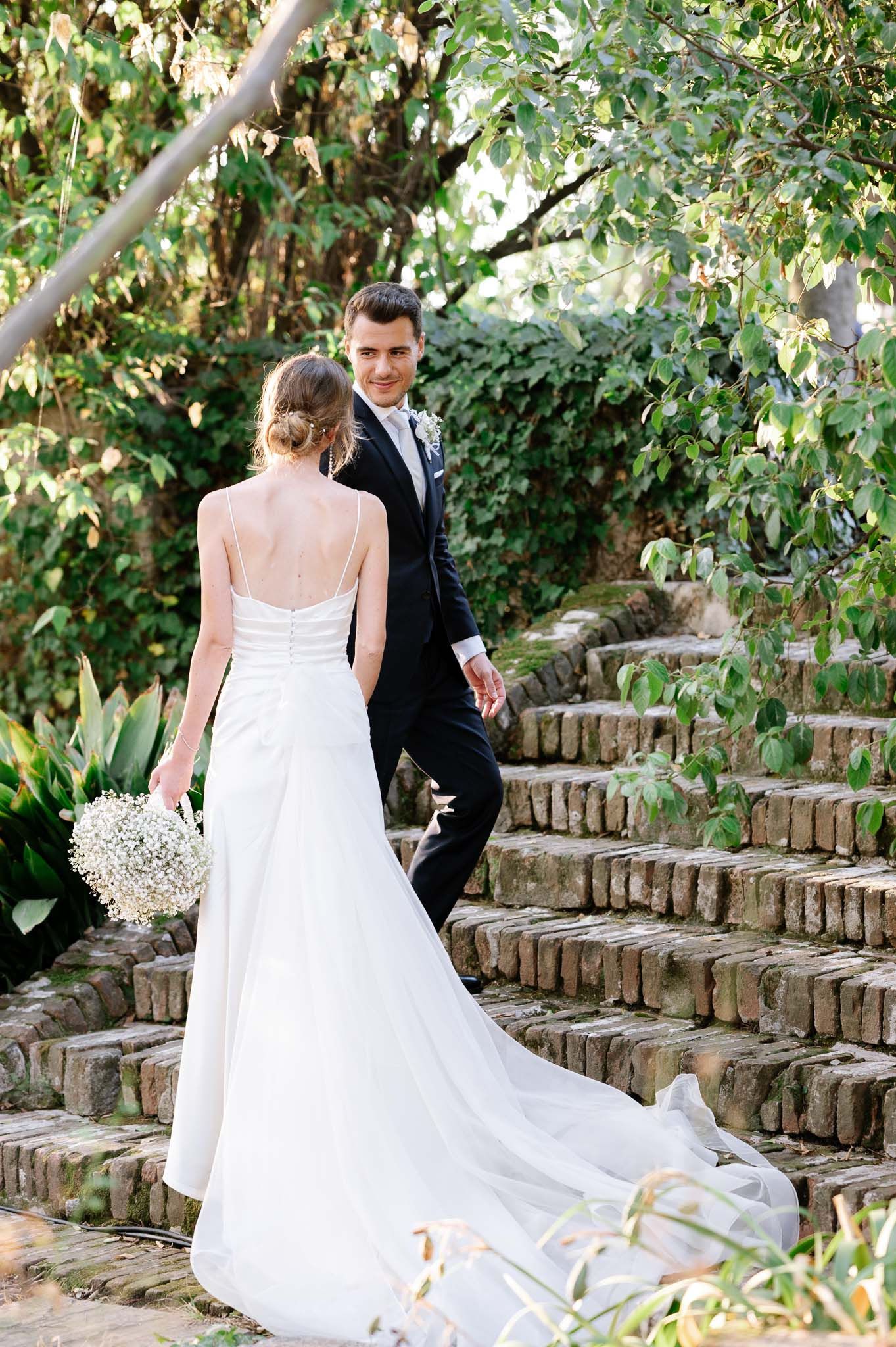 Bride in flowing white dress descends stone garden steps with groom following.