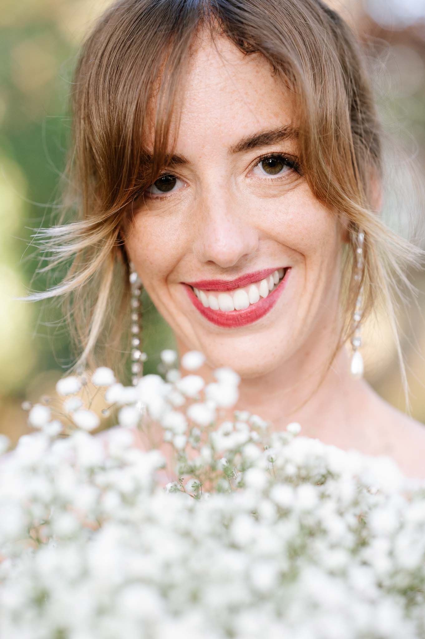 Radiant bride with classic red lipstick holding white baby's breath bouquet against outdoor setting.