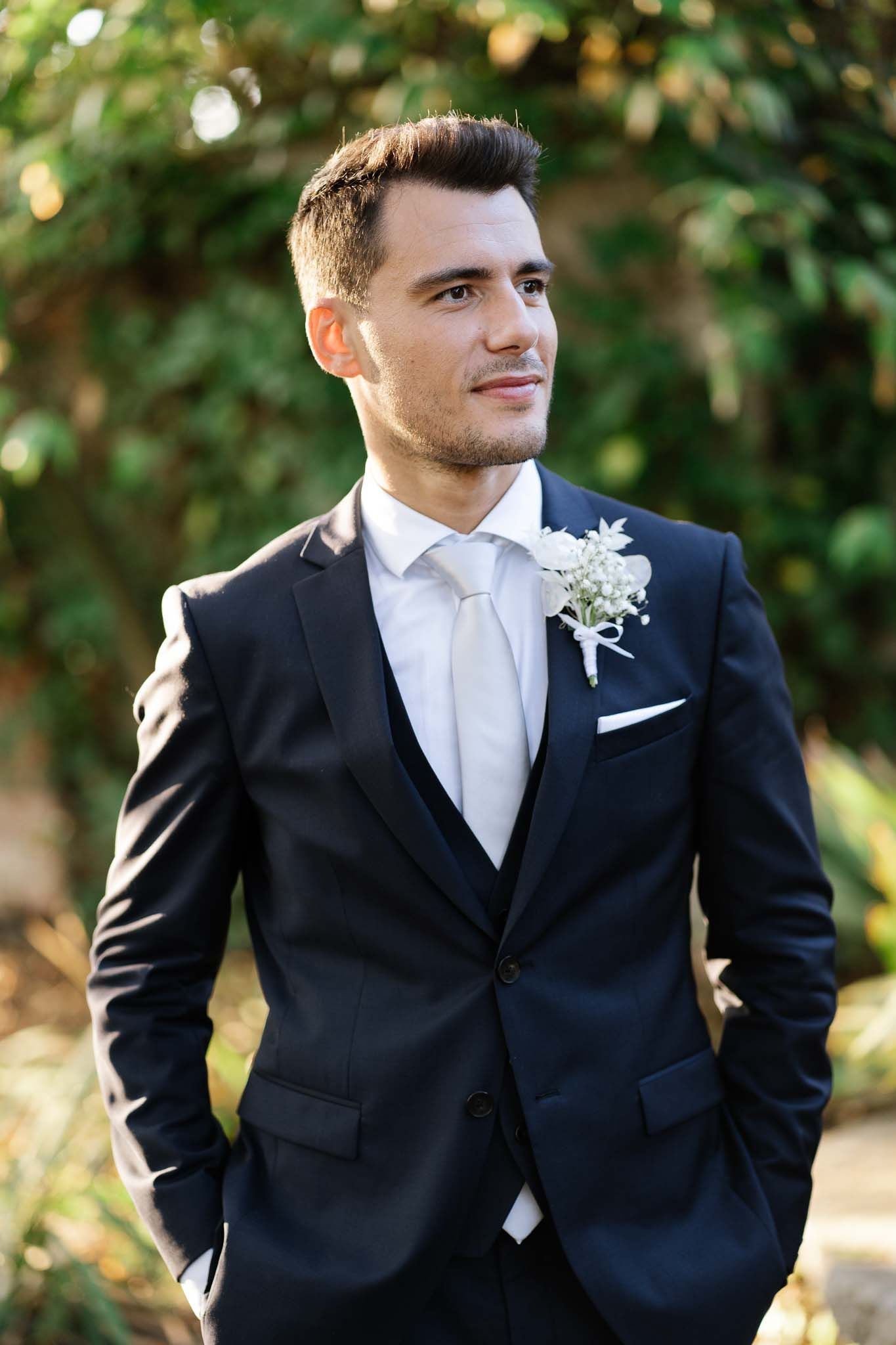 Elegant groom in classic black tuxedo with white boutonniere against garden background.