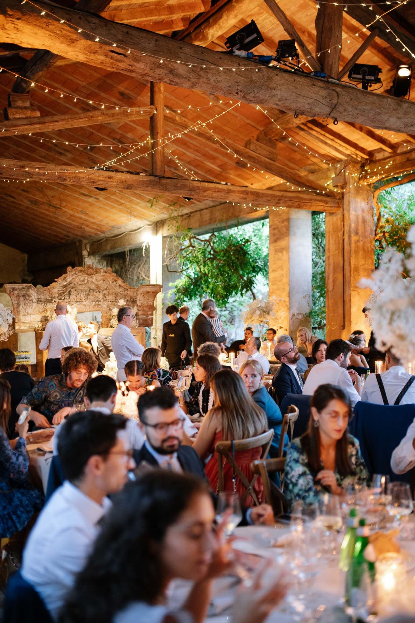 Wedding reception guests dining under rustic wooden beams with string lights in an intimate outdoor venue setting.
