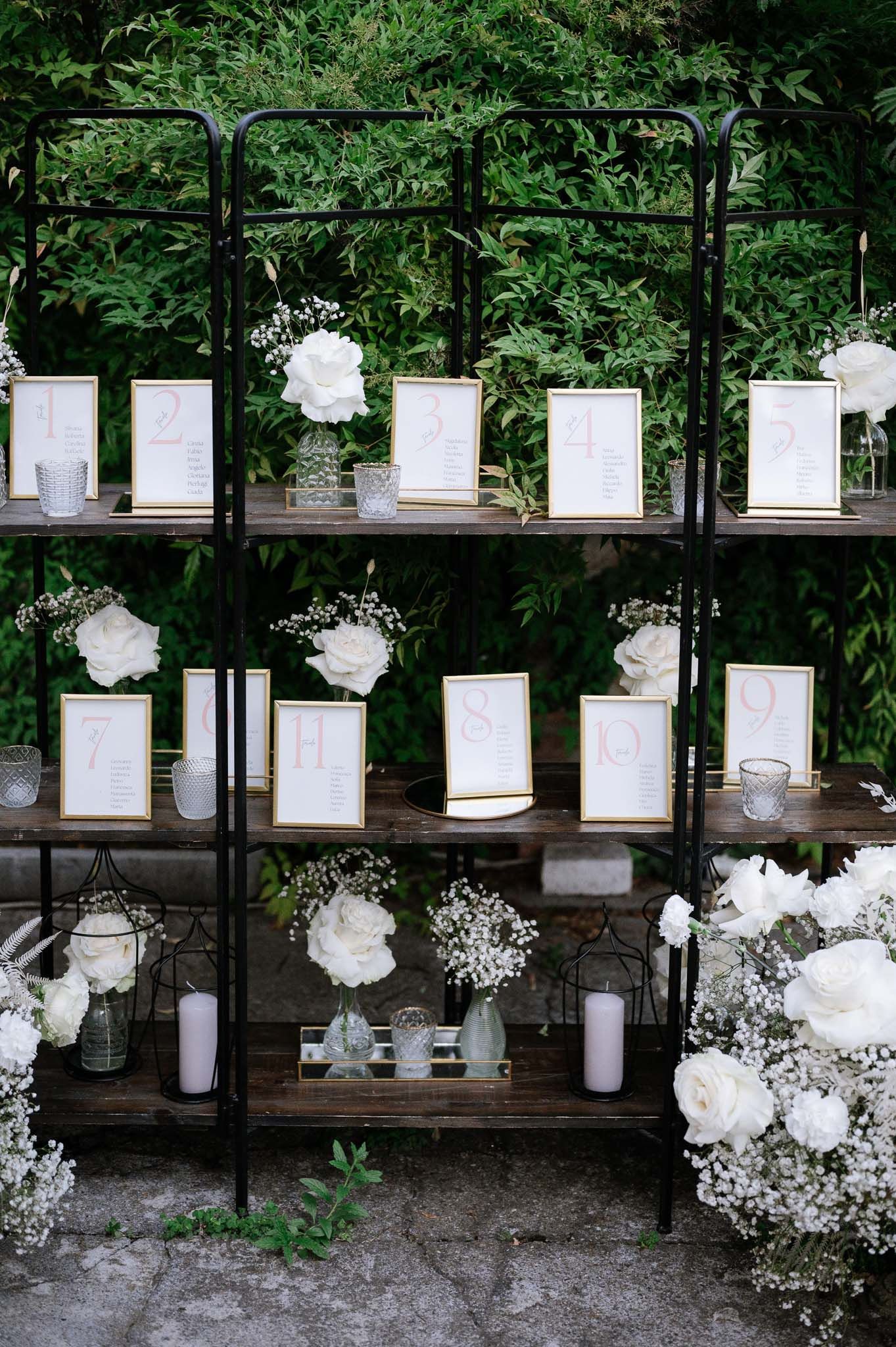 Elegant wedding seating chart display with white floral arrangements on black metal shelving unit.