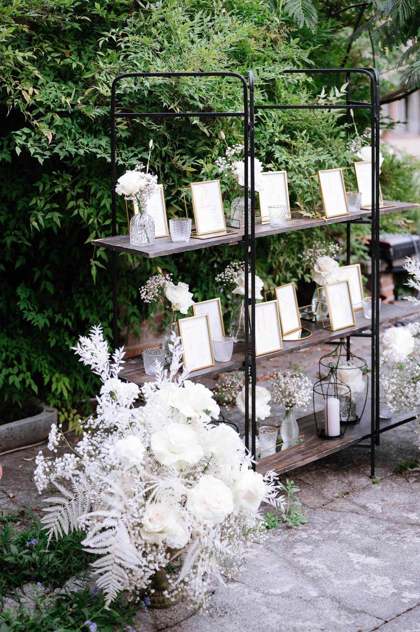 Wedding seating chart displayed on black metal shelves decorated with white flowers and candles.