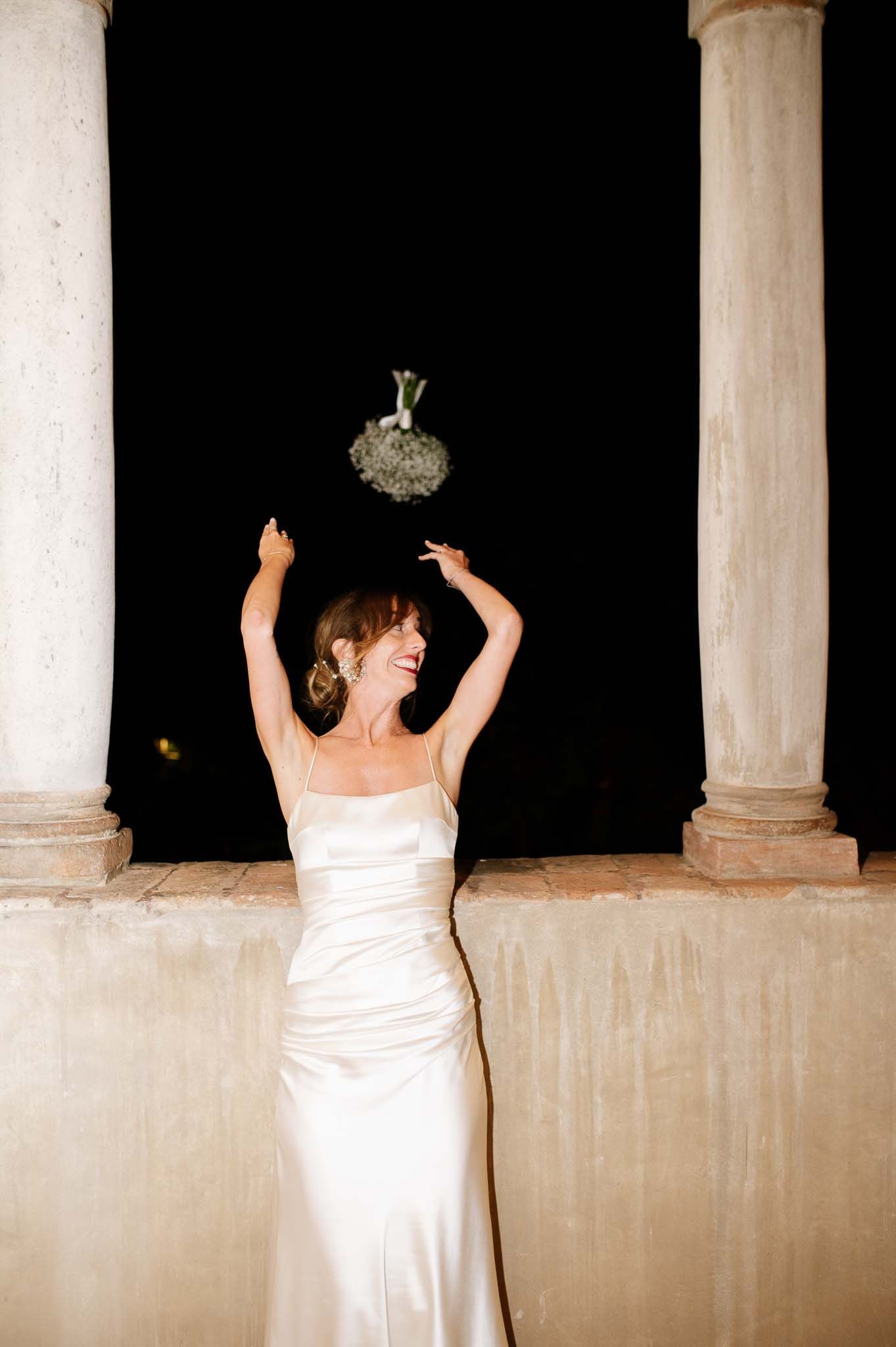 A bride in white dress tosses her bouquet between elegant columns at night.