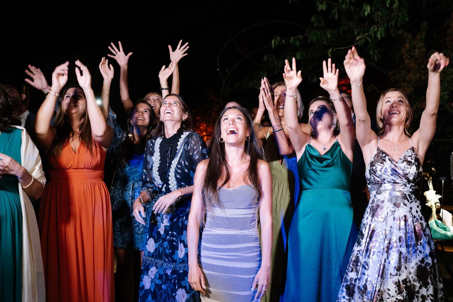 Wedding guests in colorful dresses reach up excitedly during reception festivities.