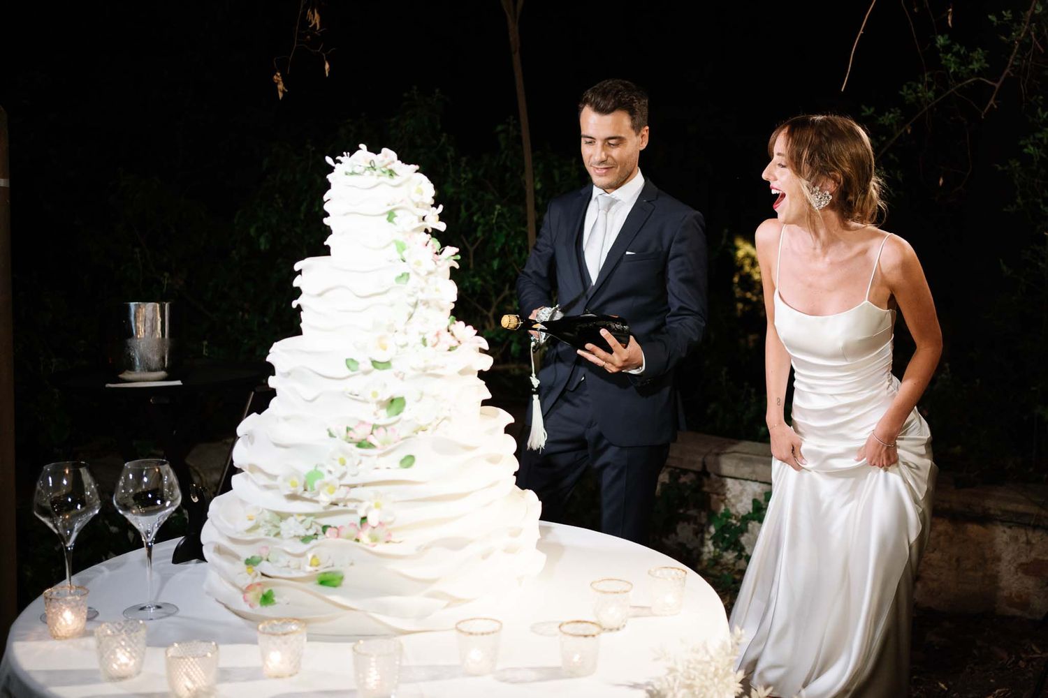 A wedding couple stands beside their elegant white tiered cake during an evening reception.