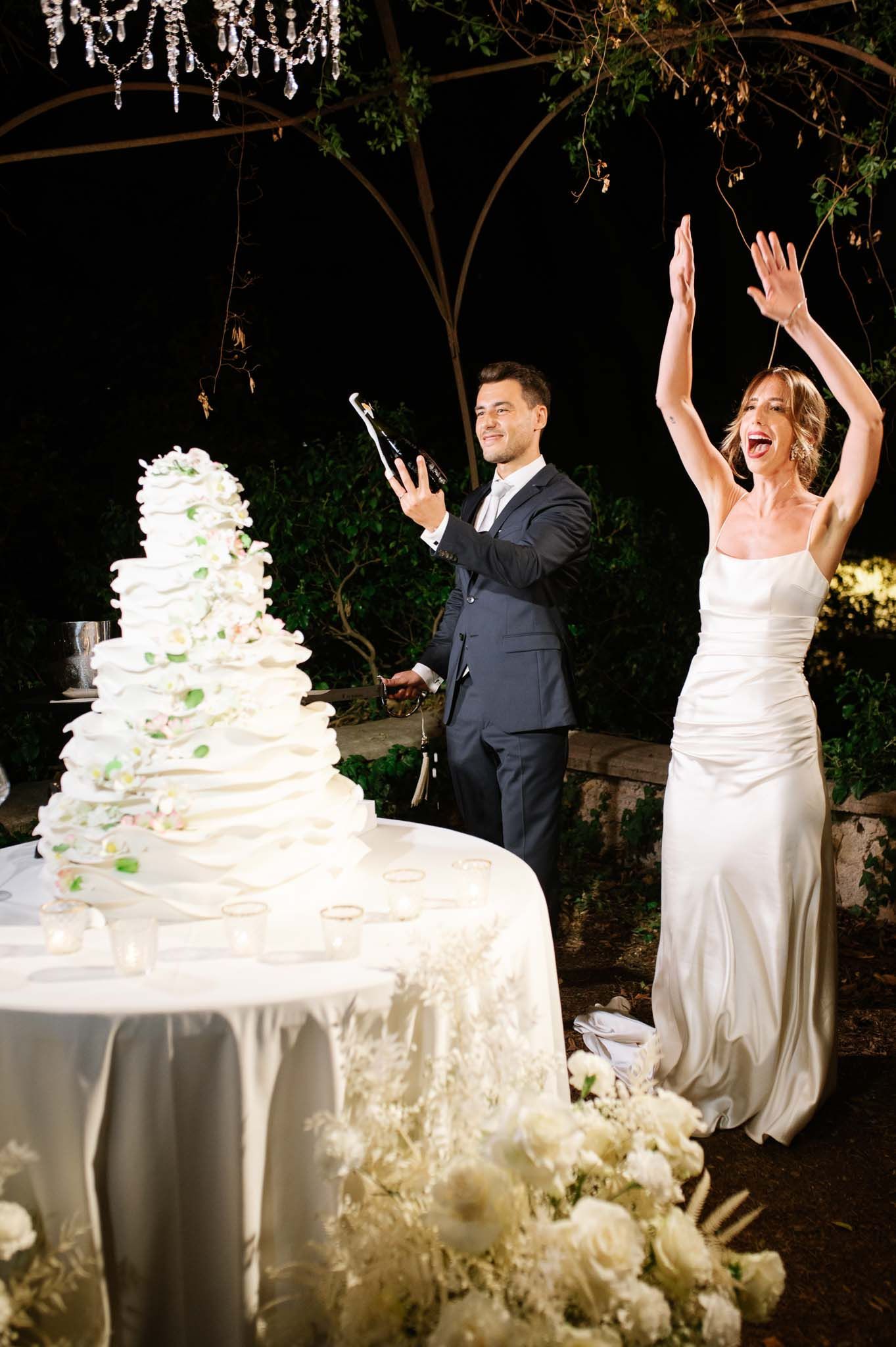 A joyful celebration moment beside an elegant white tiered wedding cake under crystal chandeliers.