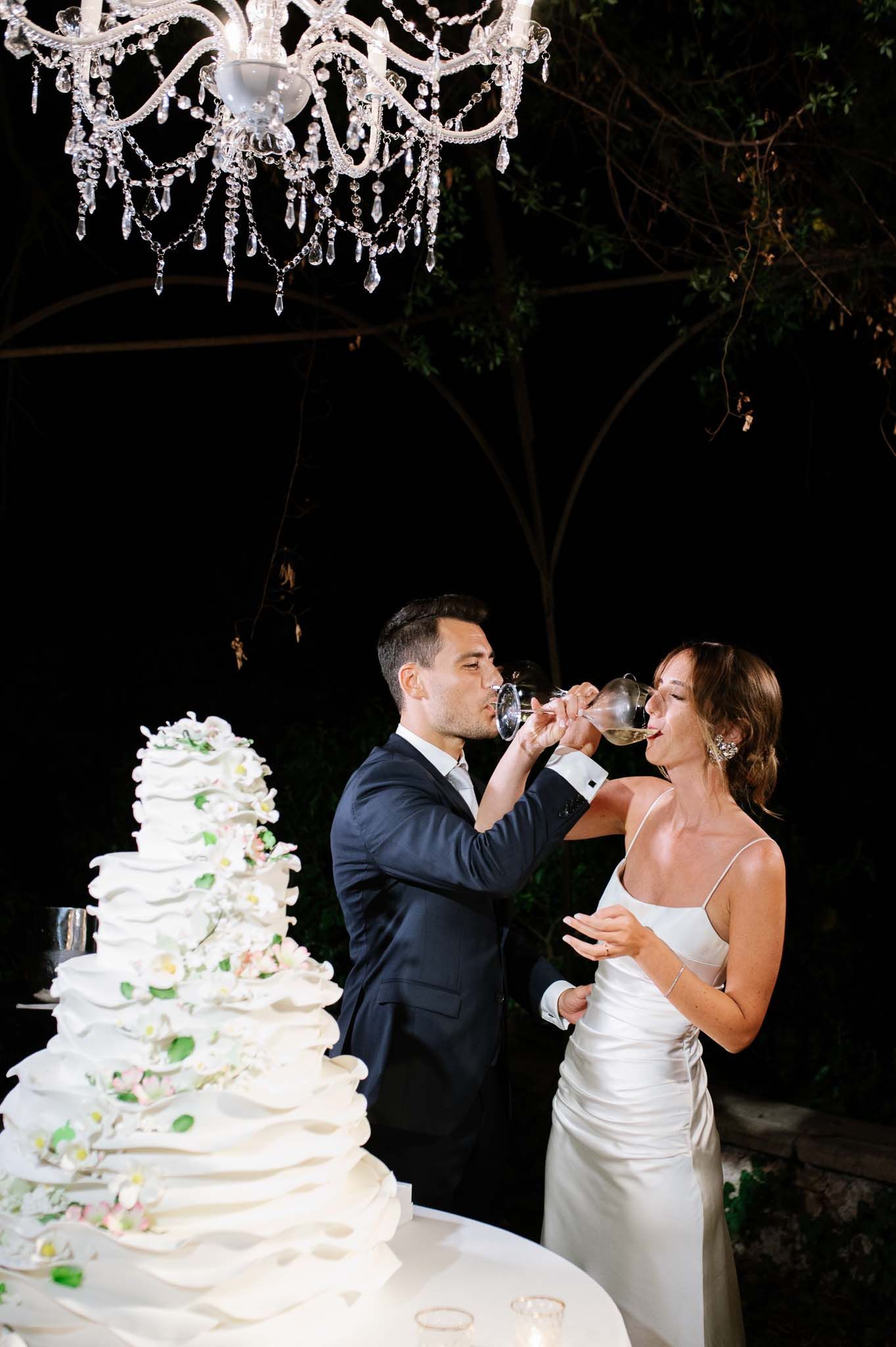 A champagne toast moment beside an elaborate white wedding cake under crystal chandeliers.