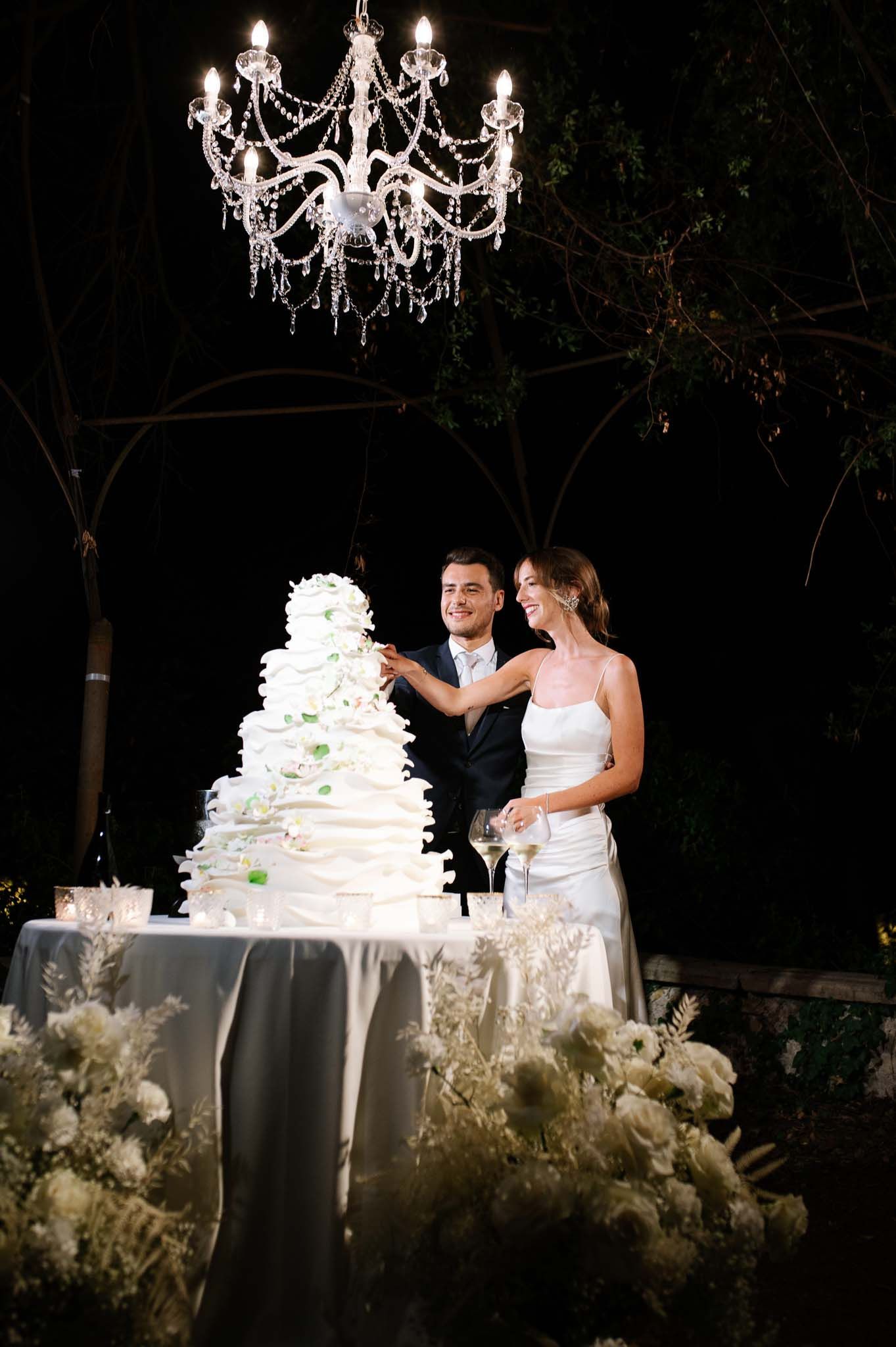 A romantic cake cutting moment under an elegant crystal chandelier at night.