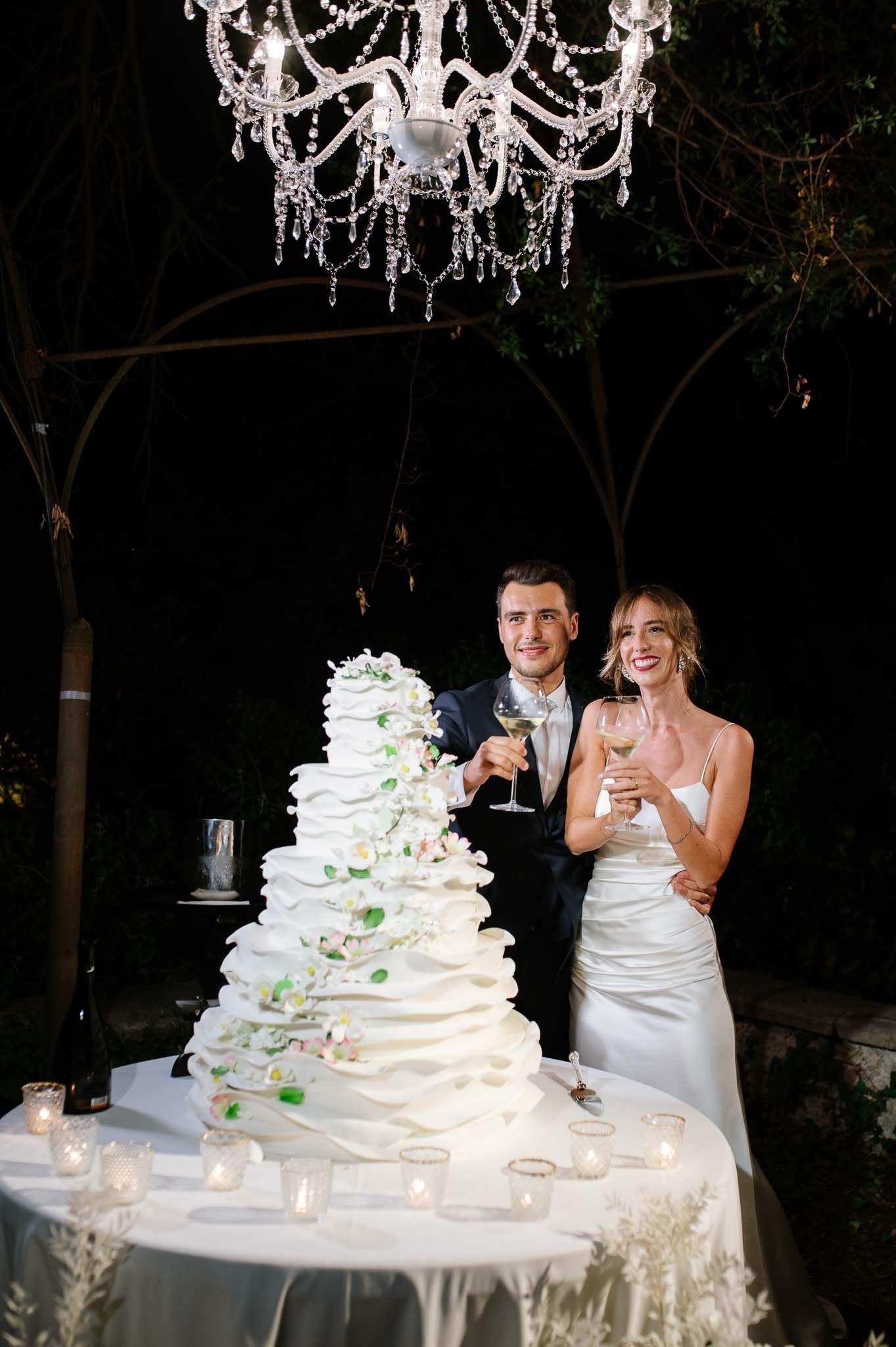 A celebratory toast beside an elegant white tiered wedding cake under crystal lighting.