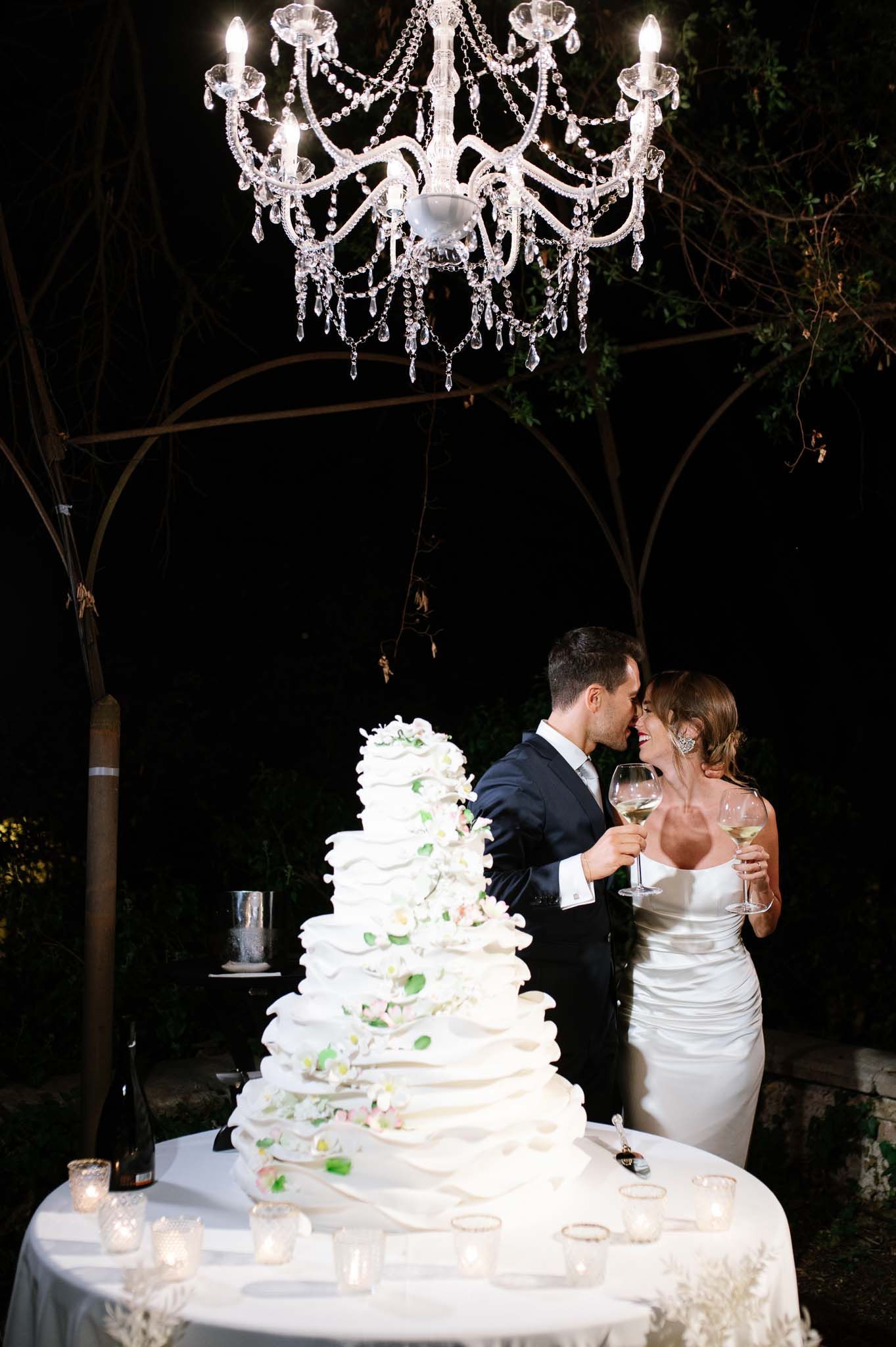 A lavish white wedding cake sits beneath an elegant crystal chandelier during an evening celebration.