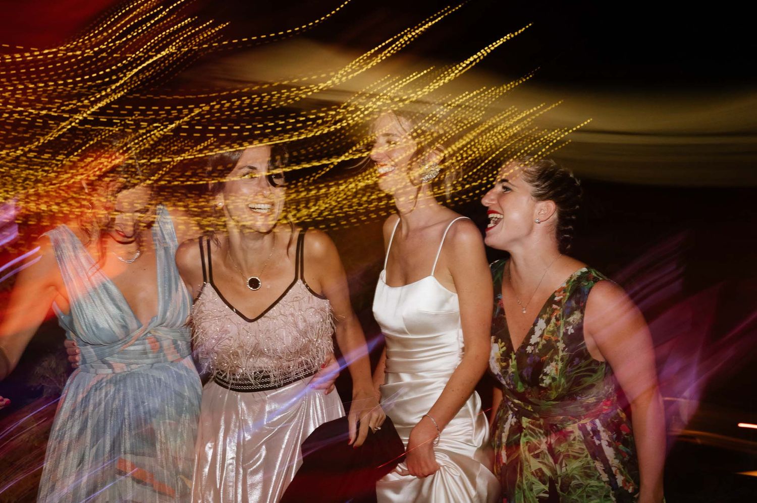 Laughing women in formal dresses dancing together at a party with colorful light trails.