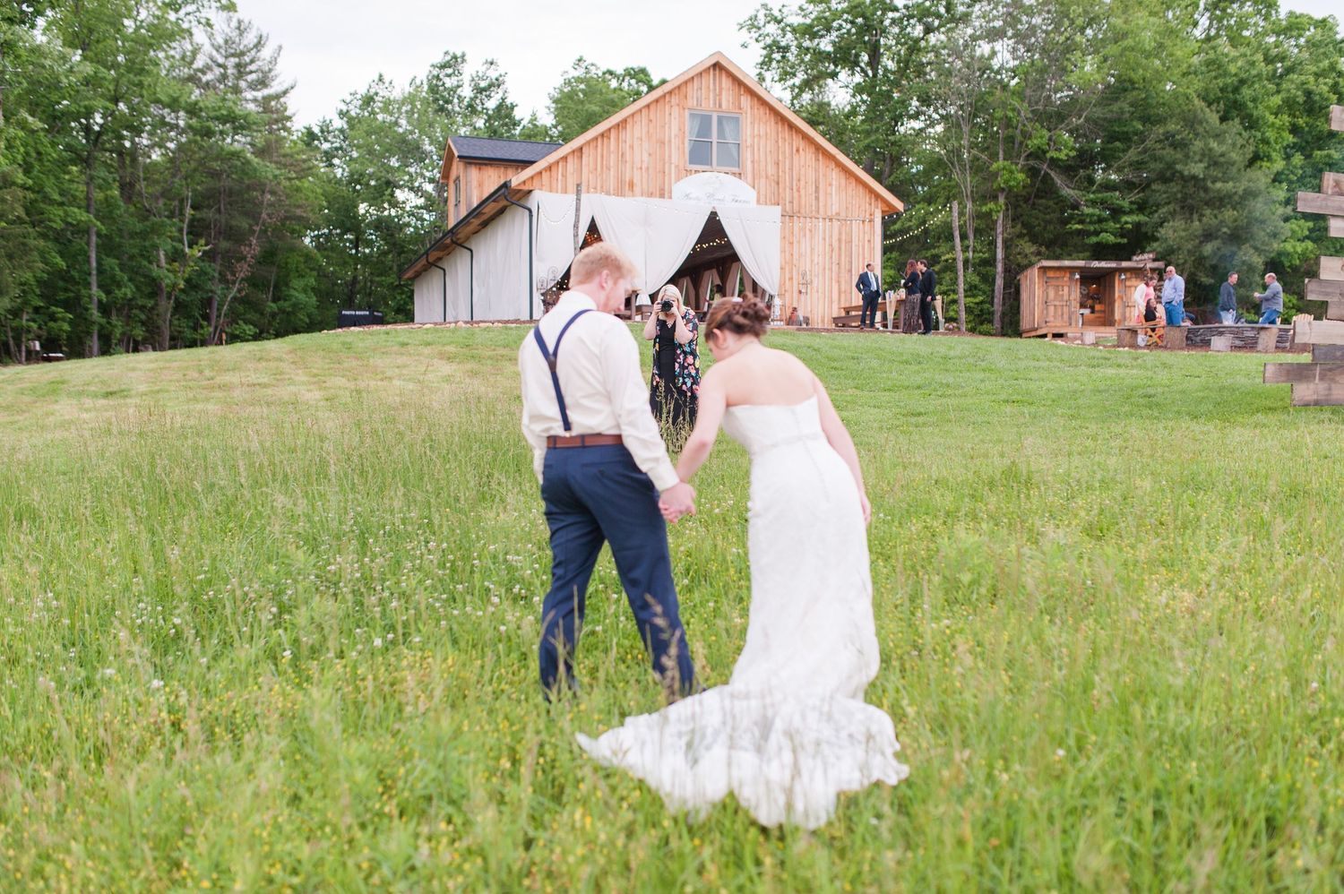 Couple walking through a sunny meadow with a rustic red barn in the background.
