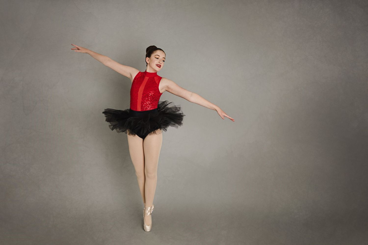 Graceful ballet dancer in red top and black tutu performing arabesque pose against grey background.