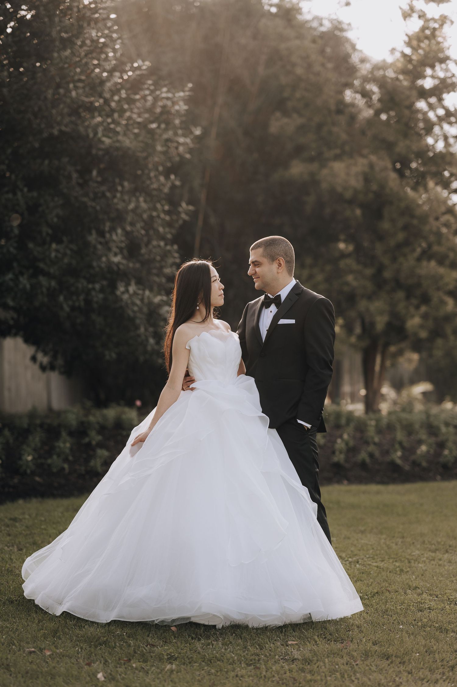 Romantic wedding portrait of couple in formal attire standing on manicured lawn at sunset.