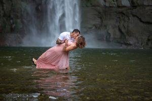 A romantic couple embraces in shallow water with a majestic waterfall cascading in the background.