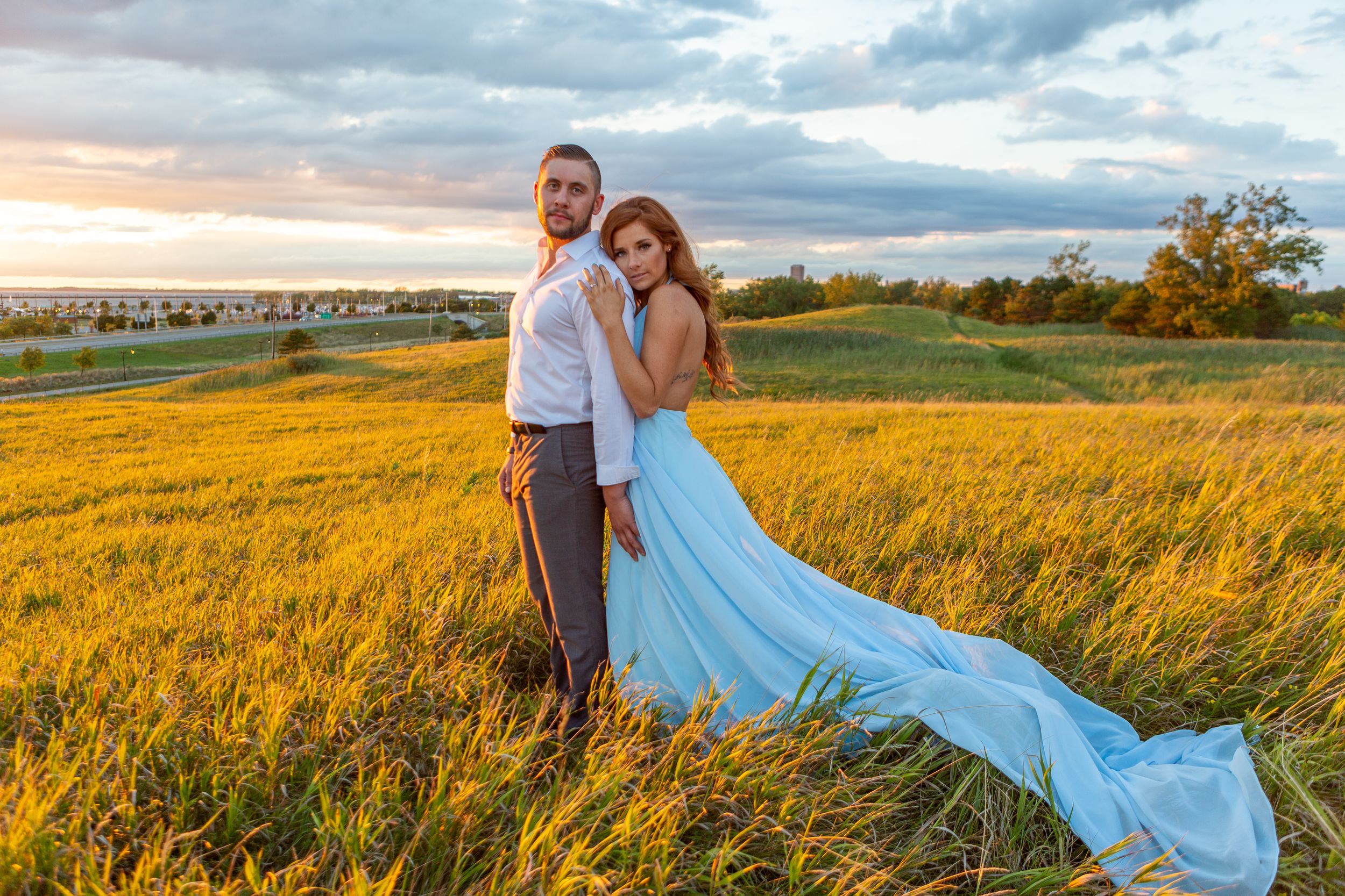 A couple in wedding attire stands in a golden field with a flowing blue dress train.