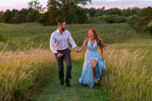 A couple runs playfully through a tall grass meadow at sunset, with her flowing blue dress catching the evening light.