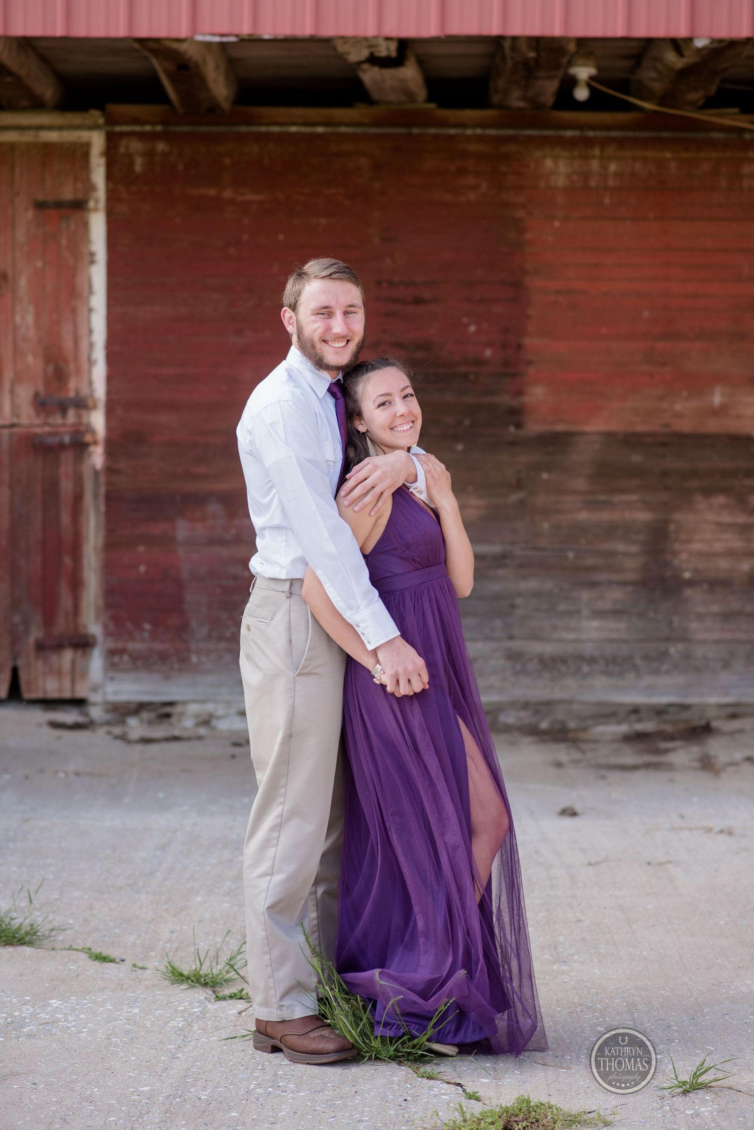 Couple poses against red barn wall, woman wearing elegant purple gown.