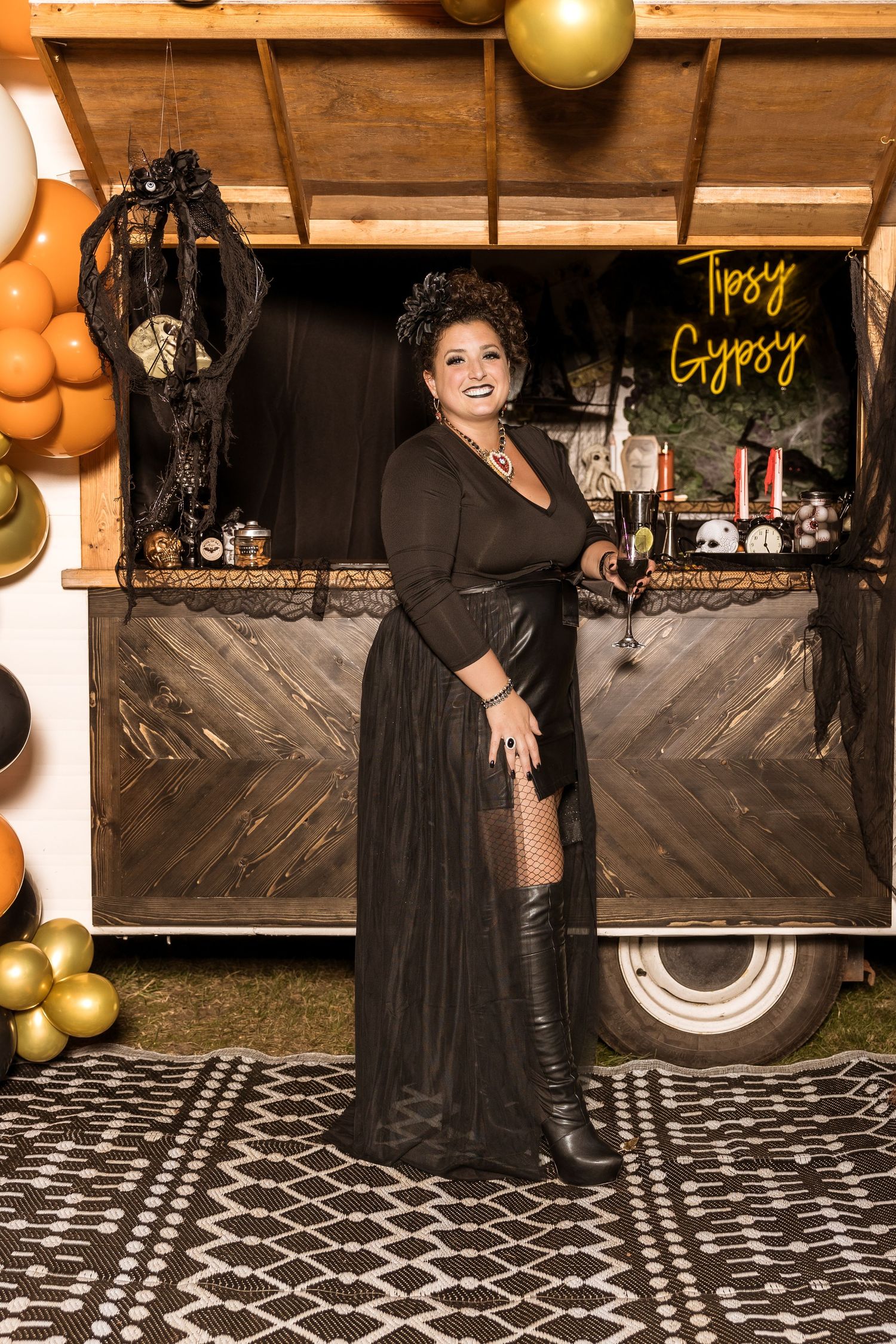 Person in long black dress poses by vintage trailer with Halloween decorations and balloons.