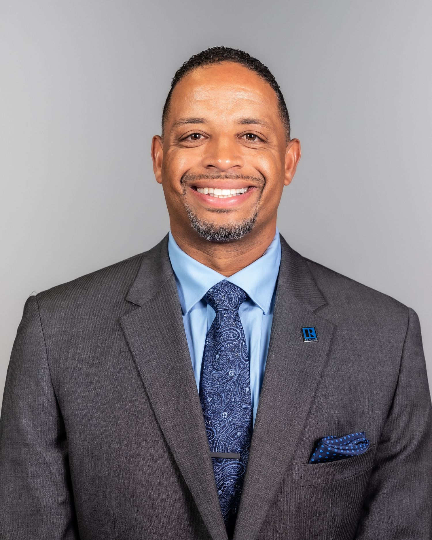 A professional headshot of a businessman wearing a grey suit with blue tie and pocket square.