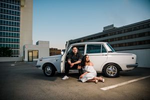 Couple sitting by classic white car in urban parking lot during sunset.