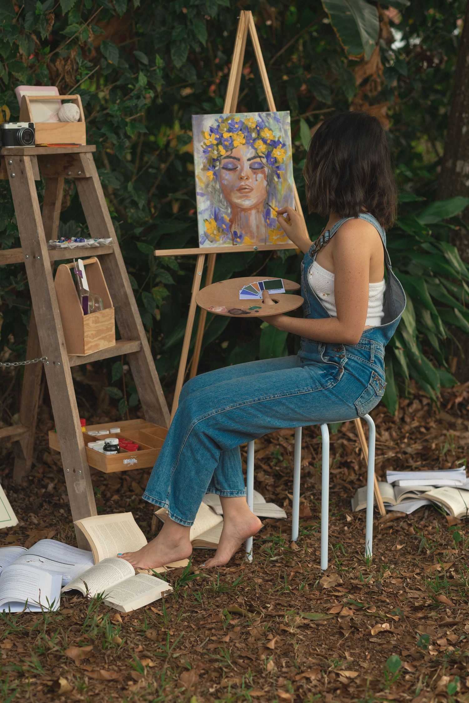 Artist painting a colorful portrait on an easel outdoors with art supplies nearby.