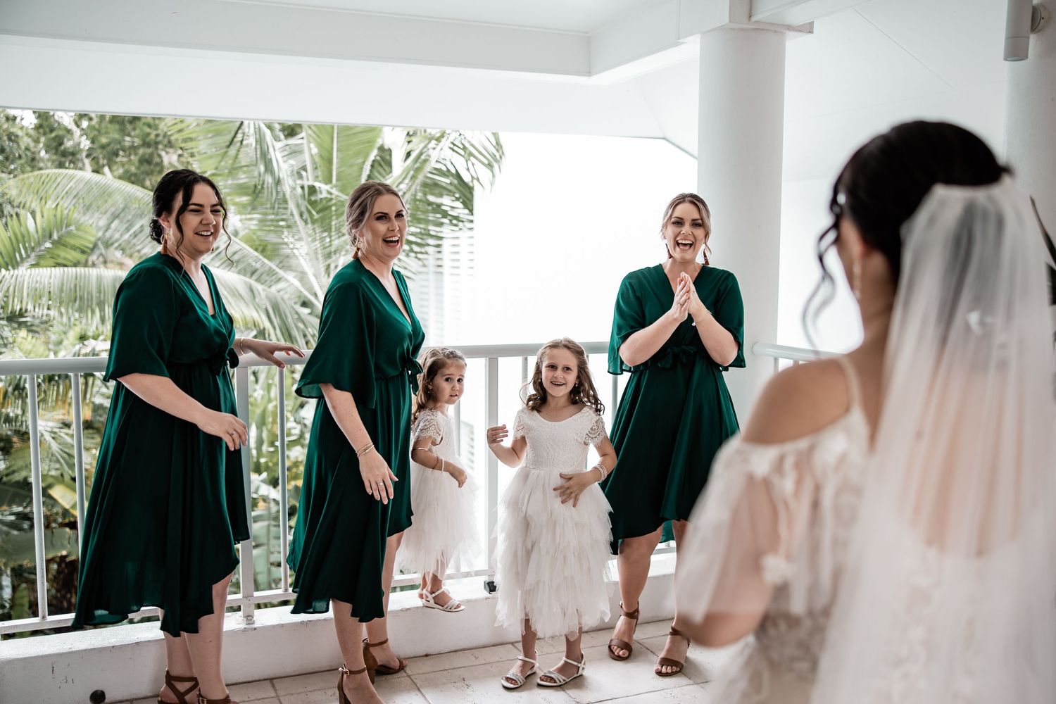 Bridesmaids in emerald green dresses react joyfully while watching the bride in her white gown and veil.