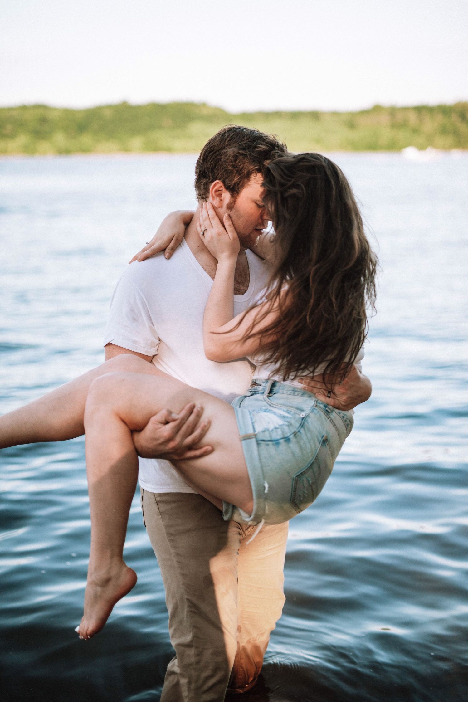 Romantic couple embracing in shallow water at sunset near a scenic shoreline.