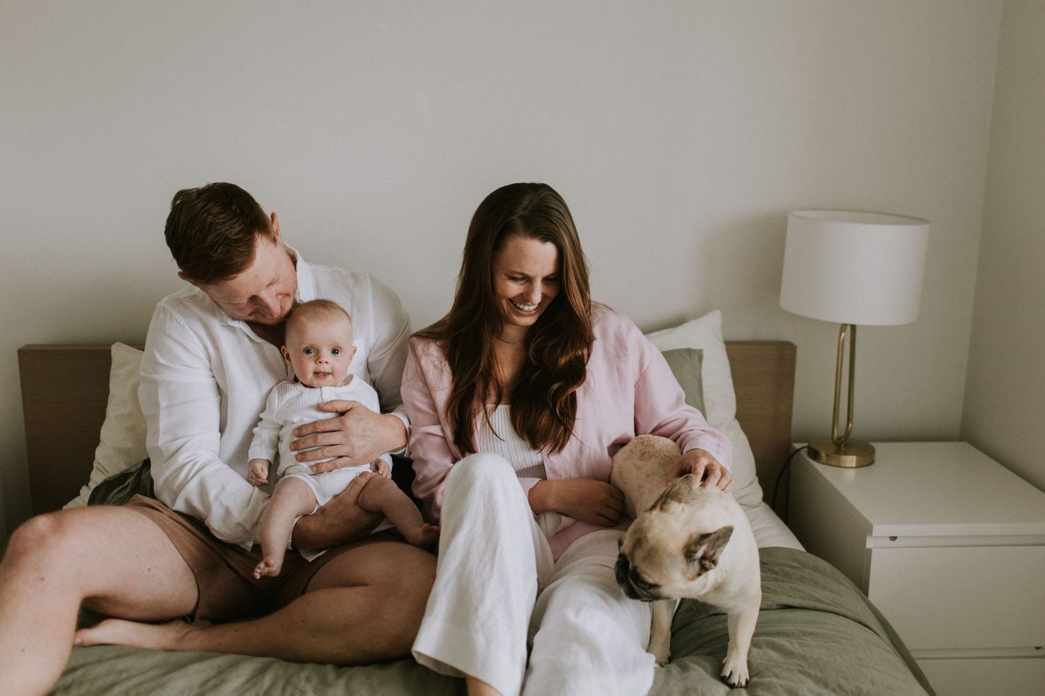 Happy couple with baby and French bulldog relaxing together on a bed in a cozy bedroom.