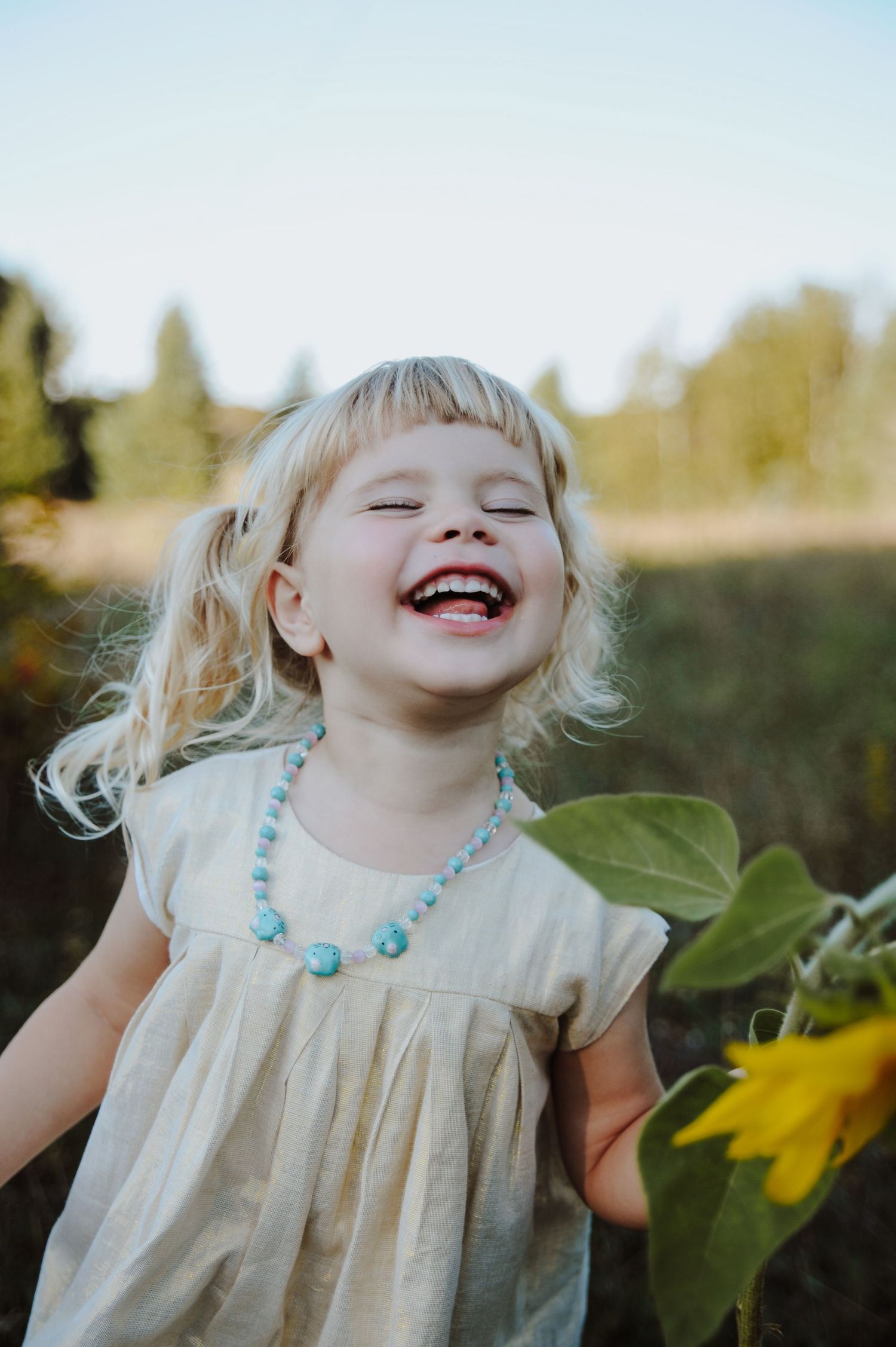 A young child in a white dress and turquoise necklace laughs joyfully near sunflowers during golden hour.